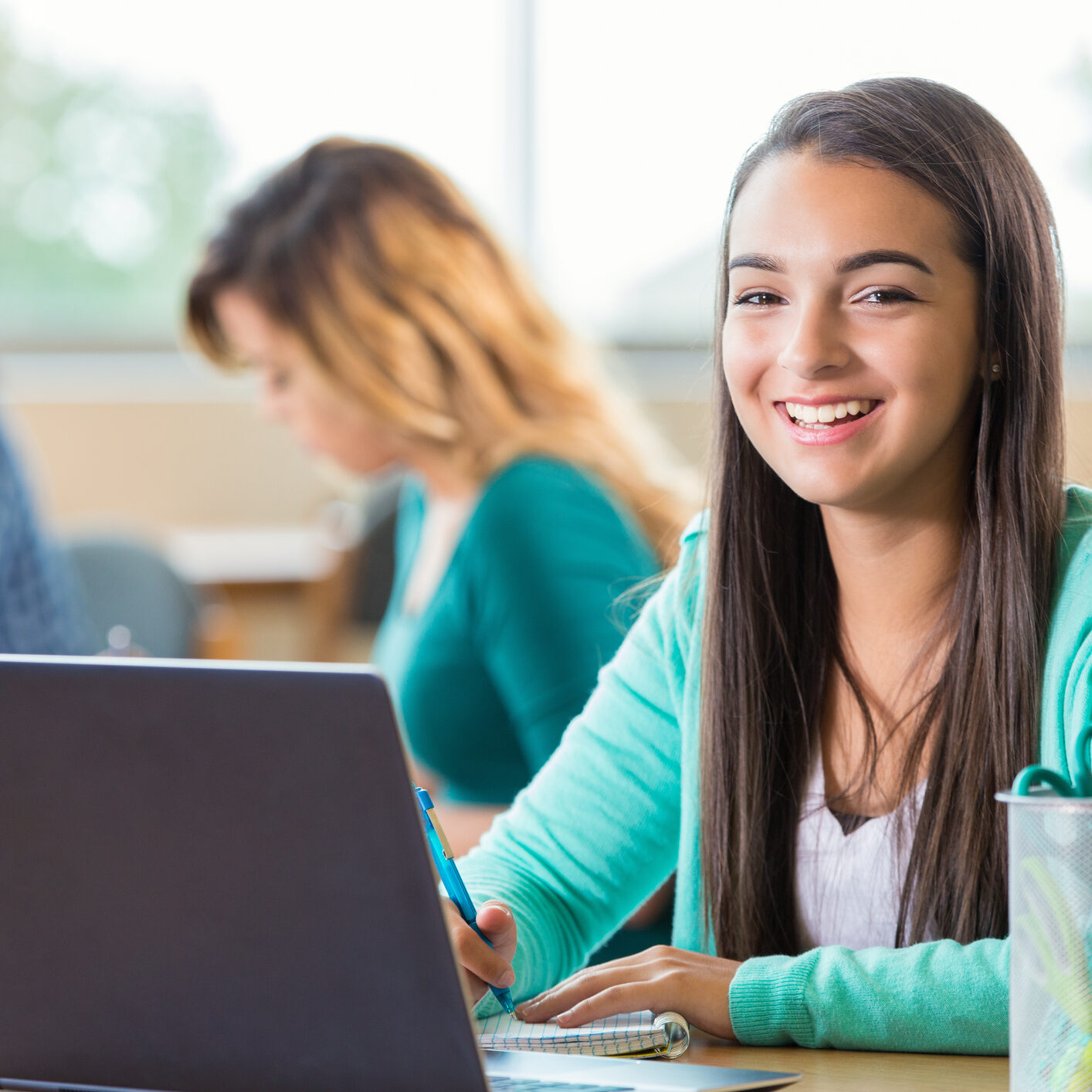 Pretty Hispanic high school student works on project in graphic design class. She is looking at the camera and smilng as she takes notes. A laptop is open on the table and art supplies are also on the table. Students are working in the background.