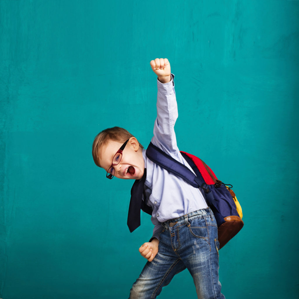 Cheerful smiling little boy with big backpack jumping and having fun against blue wall. Looking at camera. School concept. Back to School